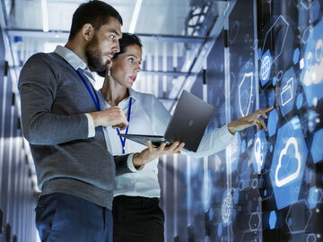 A male IT specialist holds a laptop and discusses work with Microsoft Azure, a service from T Business, with a female server technician.