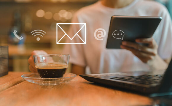 A women sitting in a cafe with a laptop on the table, a coffee in one hand and holding tablet in the other hand, using wireless technology to work remotely.