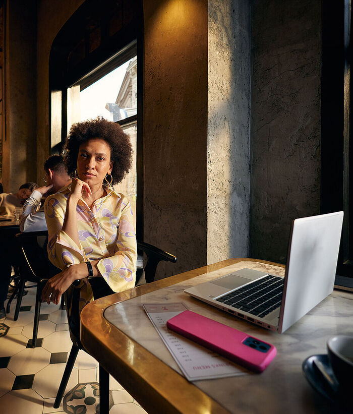 New Work A woman is sitting in a café with a modern laptop in front of her, and a smartphone lying beside it. In the background, other guests are sitting at tables.