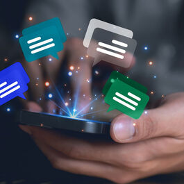 A person sitting on a desk with a laptop on it holding a smartphone in the hands and floating chat icons floating above the smartphone.