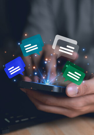 A person sitting on a desk with a laptop on it holding a smartphone in the hands and floating chat icons floating above the smartphone.
