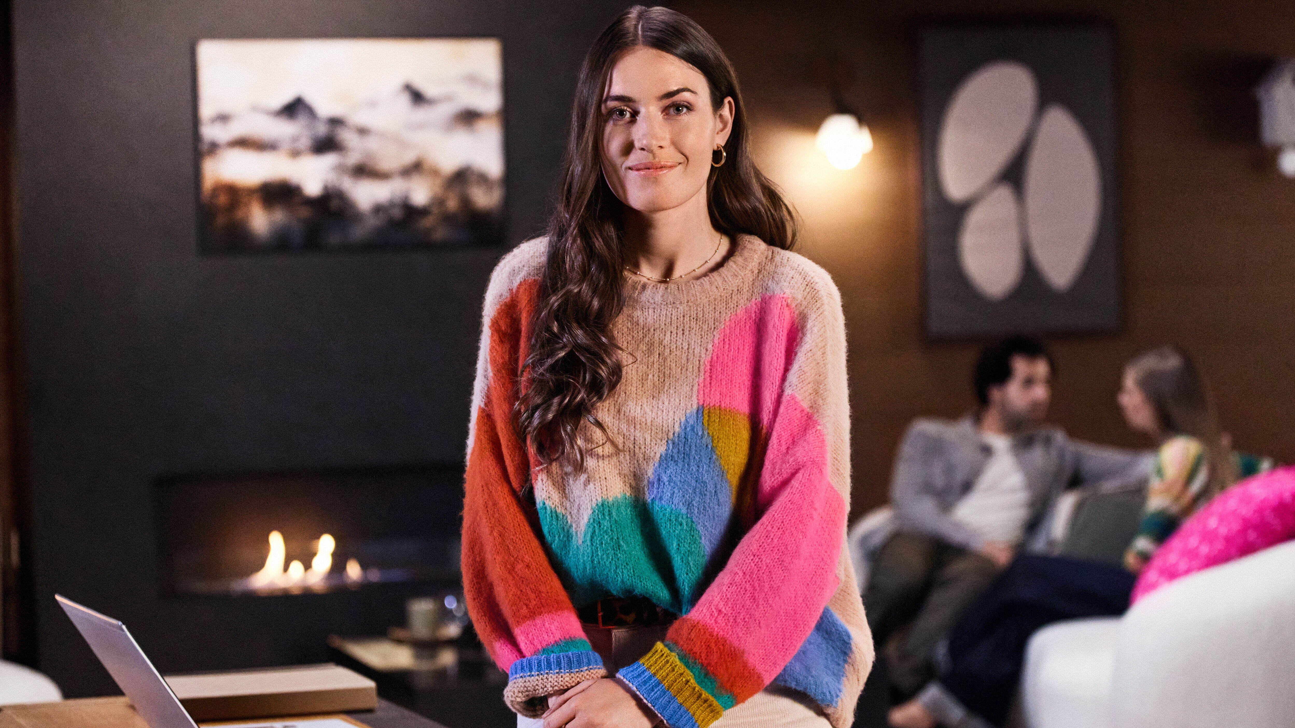 Remote work solutions A young business woman is leaning against her desk in her office with a laptop on her desk, an open fireplace in the background and two people chatting on a sofa.