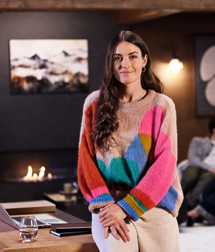 Remote work solutions A young business woman is leaning against her desk in her office with a laptop on her desk, an open fireplace in the background and two people chatting on a sofa.