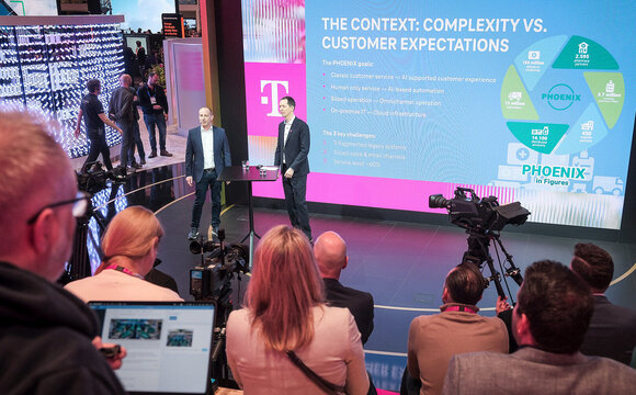 Two presenters on a stage stand before a large screen detailing Deutsche Telekom's "PHOENIX" goals and figures during a presentation at the Mobile World Congress (MWC) 2026.