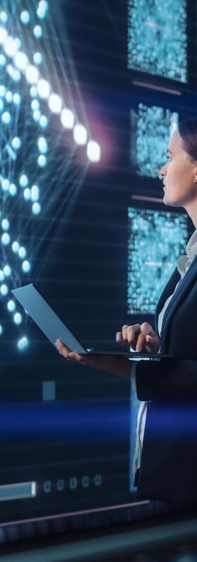 SD-X Young woman with a laptop in her hands standing in front of a big screen showing data. This shows how T Business Europe is managing the whole IT infrastructure for their clients.