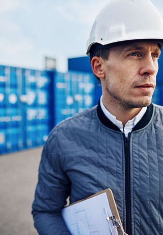 Engineer wearing a hardhat and holding a clipboard standing alone on a large commercial shipping dock, knowing everything runs smoothly because of smart solutions for smart ports from Telekom Business Europe.