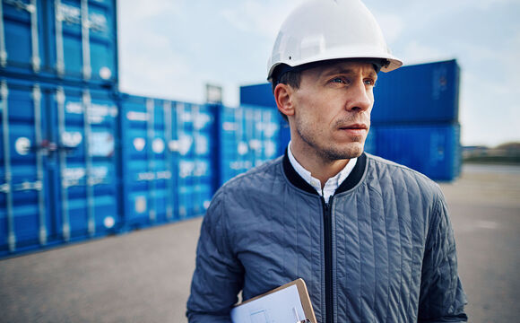 Engineer wearing a hardhat and holding a clipboard standing alone on a large commercial shipping dock, knowing everything runs smoothly because of smart solutions for smart ports from Telekom Business Europe.
