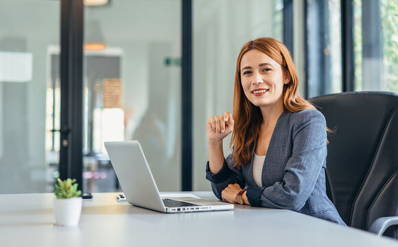 A professional woman sits in a modern office, smiling confidently at her laptop because she simplifies her communication with Microsoft Teams Phone provided by Telekom Business Europe.
