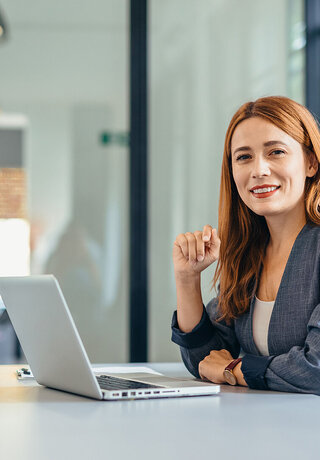 A professional woman sits in a modern office, smiling confidently at her laptop because she simplifies her communication with Microsoft Teams Phone provided by Telekom Business Europe.