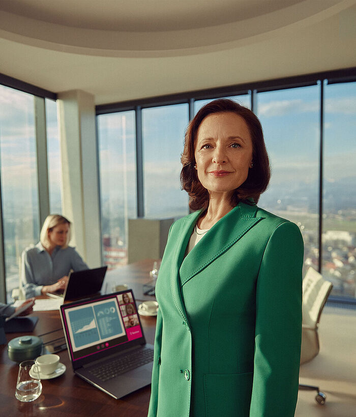 Small Business Security A businesswoman stands in a modern office in front of a desk; in the background, other colleagues can be seen participating in a video call.