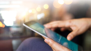 Secure Connectivity Against a blurred urban backdrop, a mobile device is held in one hand using secure connectivity solutions from T Business.