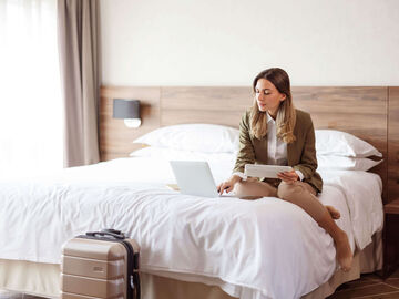 Hospitality IT Solutions A woman sitting on a bed with her laptop in a hotel, that is using the best hospitality IT solutions