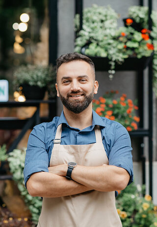 Modern business smiling confident in apron with crossed arms because he is using the digital payment solutions from T Business Europe, standing in front of his flower shop with plants in pots and sign is open.