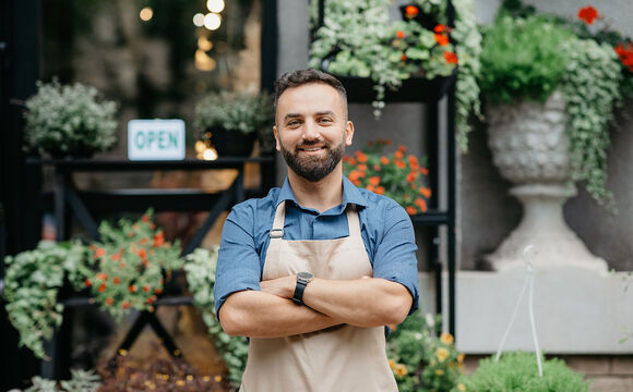 Modern business smiling confident in apron with crossed arms because he is using the digital payment solutions from T Business Europe, standing in front of his flower shop with plants in pots and sign is open.