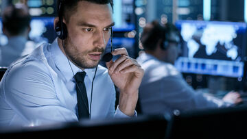 NIS2 Requirements A middle-aged man with a headset sitting in a call center in front of a screen and colleagues in the back.