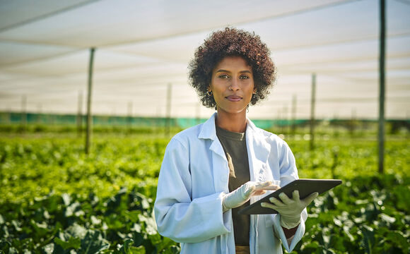 A woman with a tablet in her hands and a field of green plants, confident because she is using smart sensors for green farming from T Business Europe.