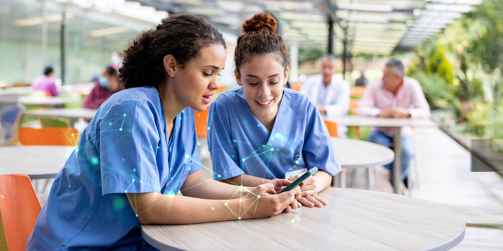 Two nurses sitting at a table with a smartphone in their hands using the 5G Campus Network provided by T Business Europe.