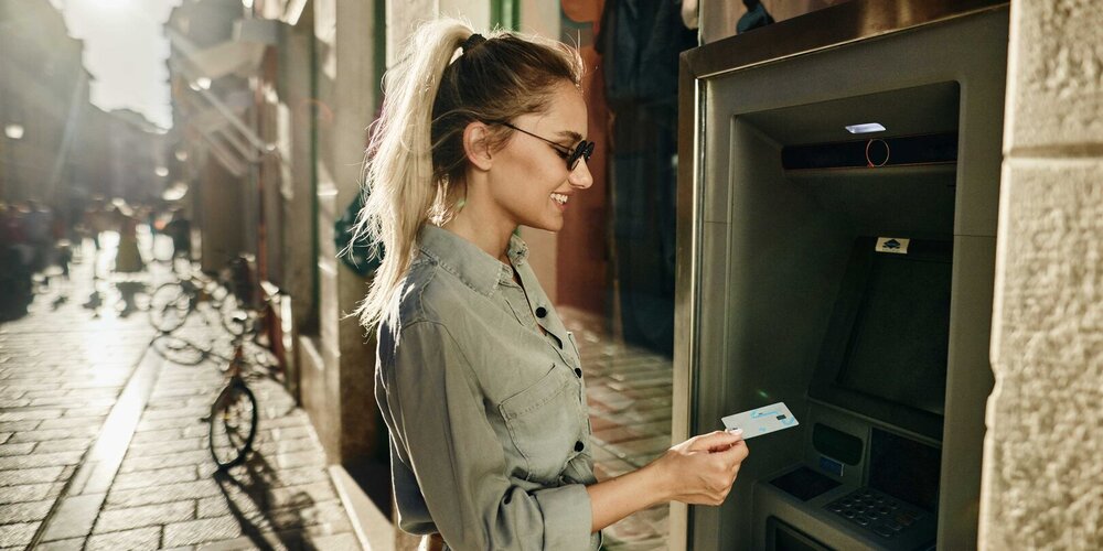 A young woman using a credit card at an outdoor ATM on a sunny street.