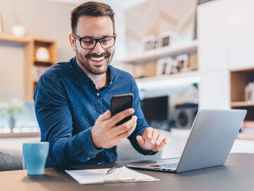 Hospitality IT Solutions A man smiling at his smartphone while searching on his laptop for hospitality IT solutions