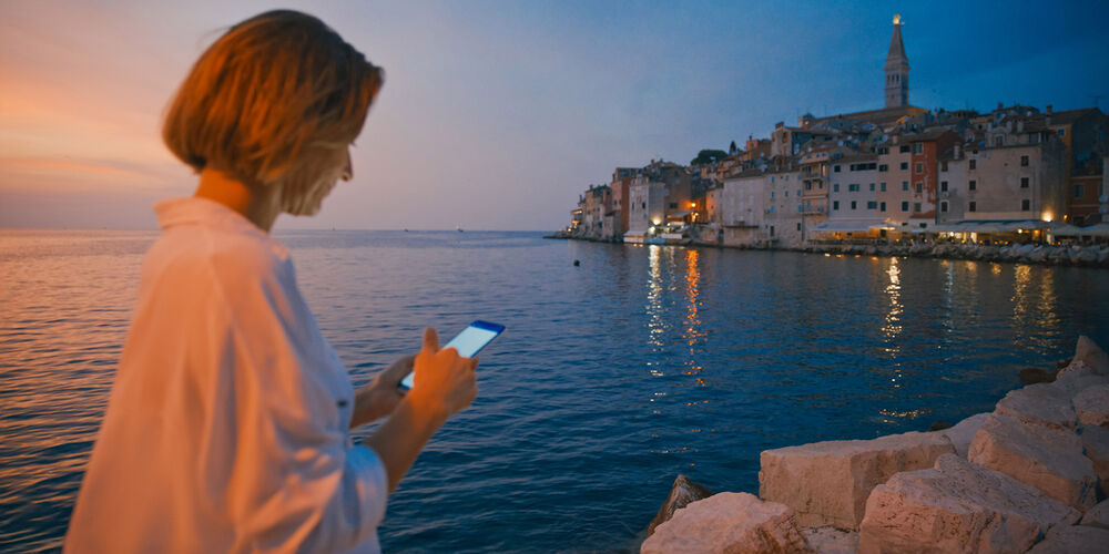 A woman with a smartphone in her hands using the OneCityApp from T Business Europe in a Croatian city with a sunset and a city in the background.