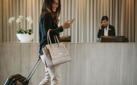 Woman using mobile phone to digitally check in via the communication platform from T Business Europe. The female business traveler walking in hotel hallway and pulling her suitcase.