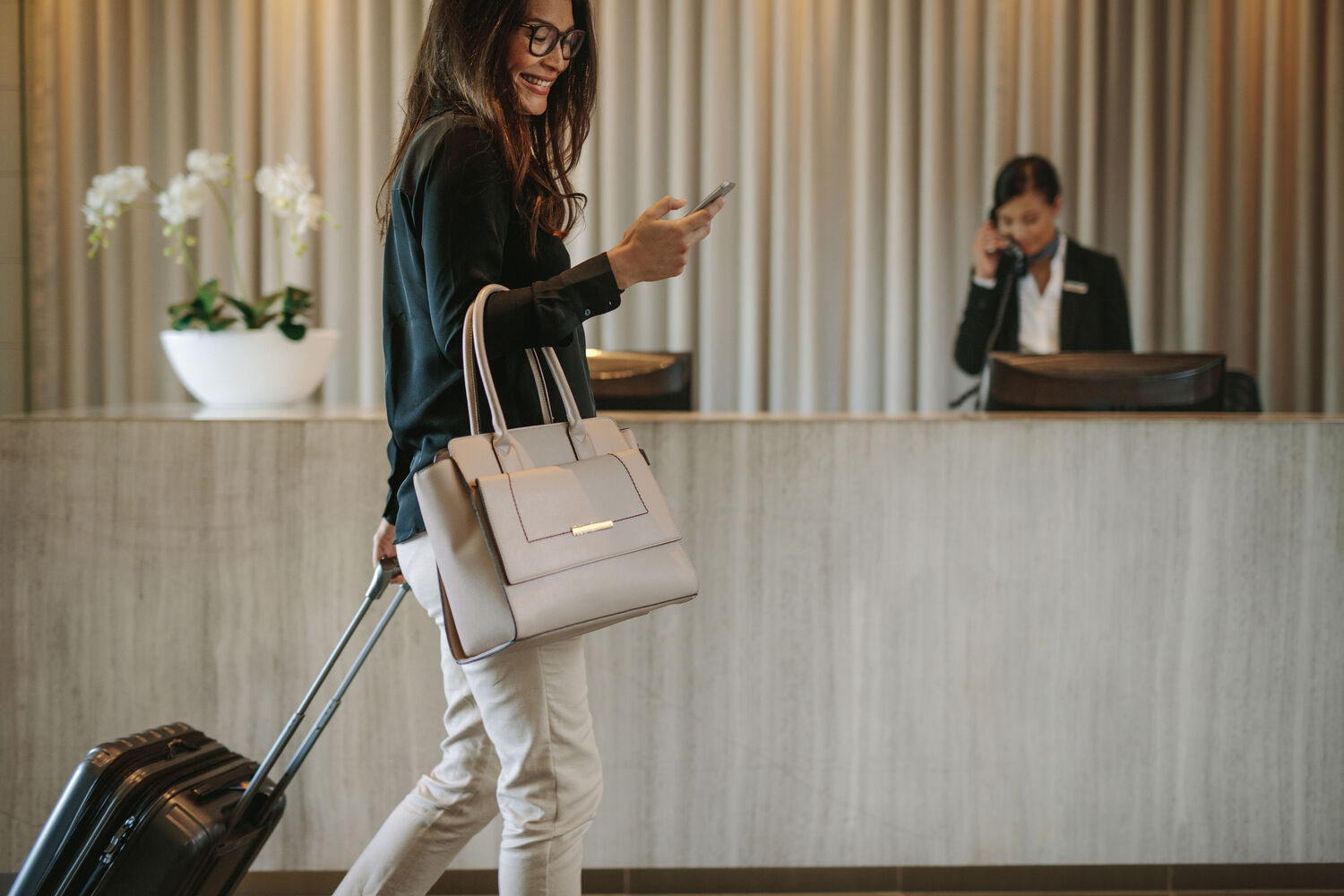 Woman using mobile phone to digitally check in via the communication platform from T Business Europe. The female business traveler walking in hotel hallway and pulling her suitcase.